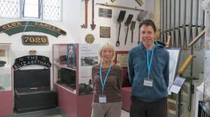 Two people stand in a museum, with exhibits and artifacts displayed in the background.