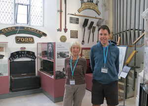 Two people stand in a museum, with exhibits and artifacts displayed in the background.